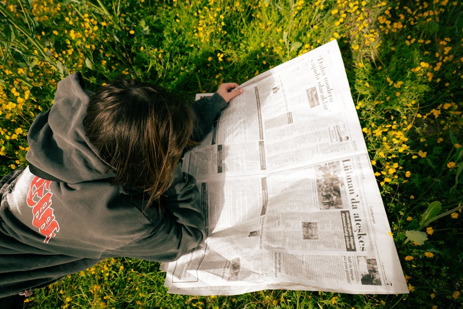 A young person enjoying reading a newspaper while lying on a flowery spring field.